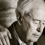 Uomo anziano affetto da Alzheimer guarda in basso e mantiene gli occhi chiusi. Ha una mano nella spalla destra dell'assistente